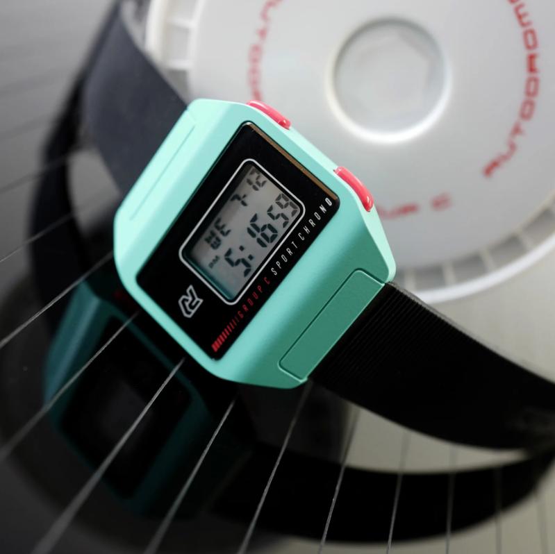 Red digital watch with a light blue strap hanging on branch with the ocean in the background